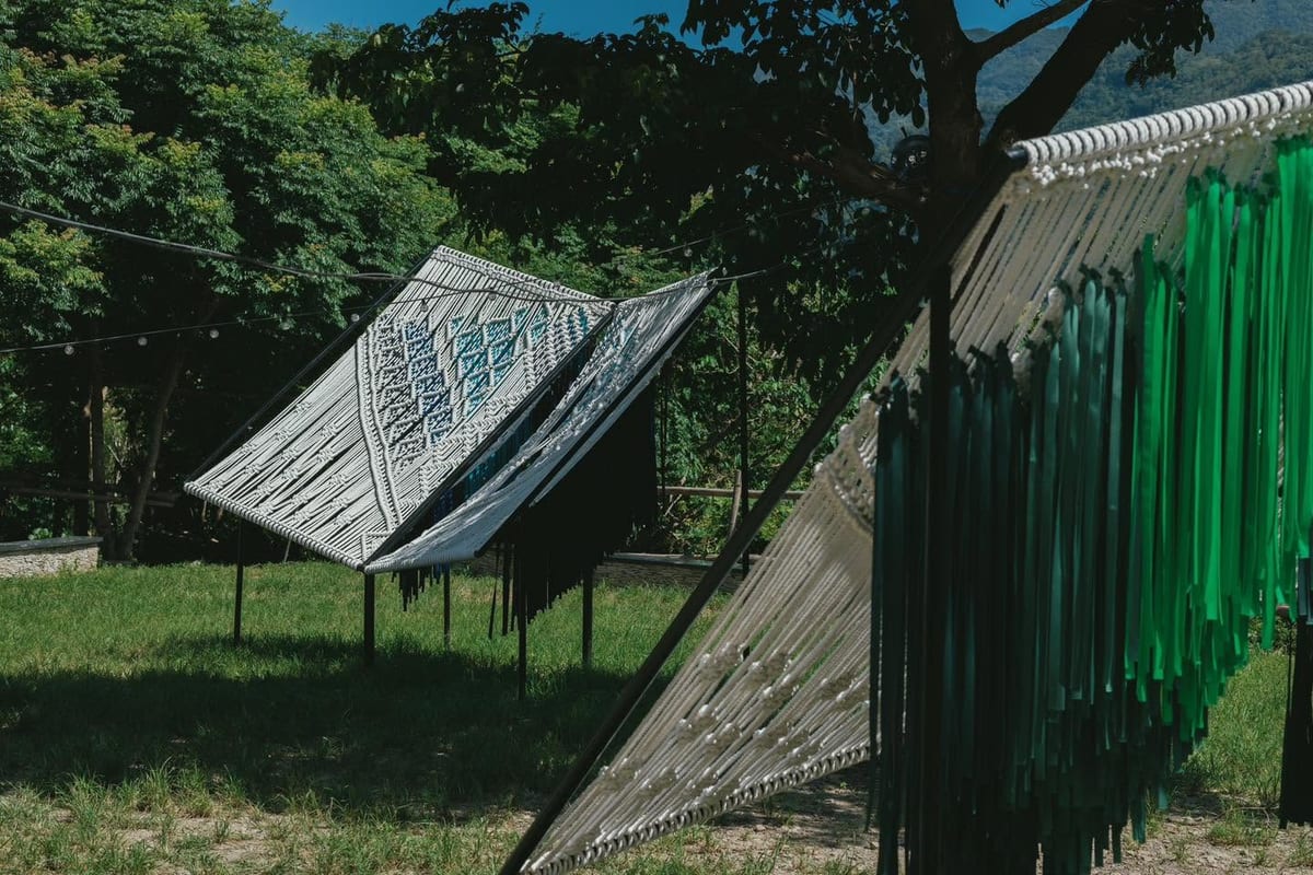 Several tilted panels of knotted cord stand on grass at the foot of a hill, their white and blue patterns casting long shadows, with a curtain of dark green fabric strips hanging in the right foreground.