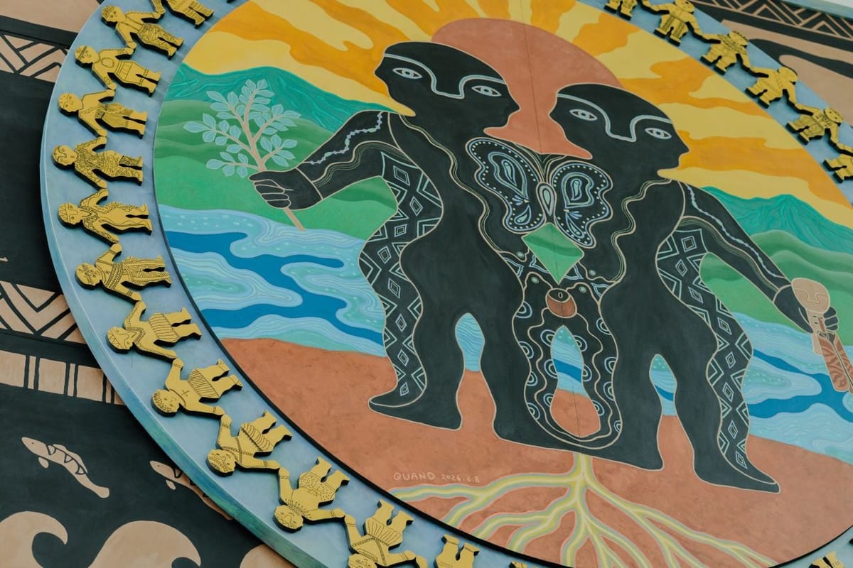 On a circular painted panel two black figures stand side by side between a rising sun, mountains, and water, their bodies patterned with Paiwan motifs; a ring of golden relief figures runs around the rim, with roots and fish below.