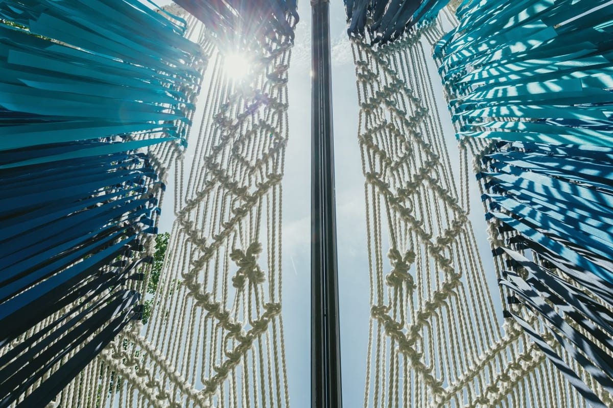 Seen from below, two symmetrical wing-like panels of knotted cream cord and blue-green fabric strips open either side of a dark upright post, with sunlight breaking through the gap.