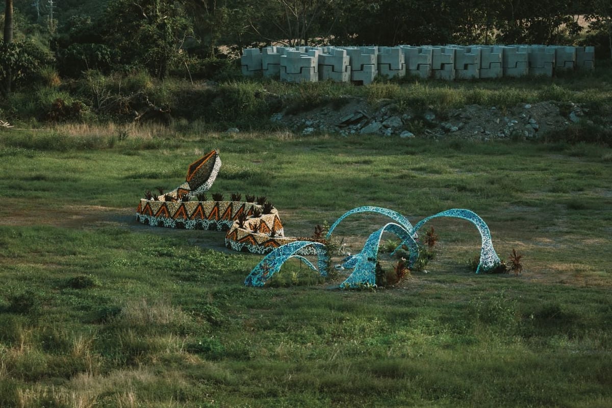 On a grassy slope at dusk, blue-green mosaic arcs curve out of the ground beside a long red-and-black patterned band, with plants growing between the arcs and a row of concrete blocks along the ridge behind.