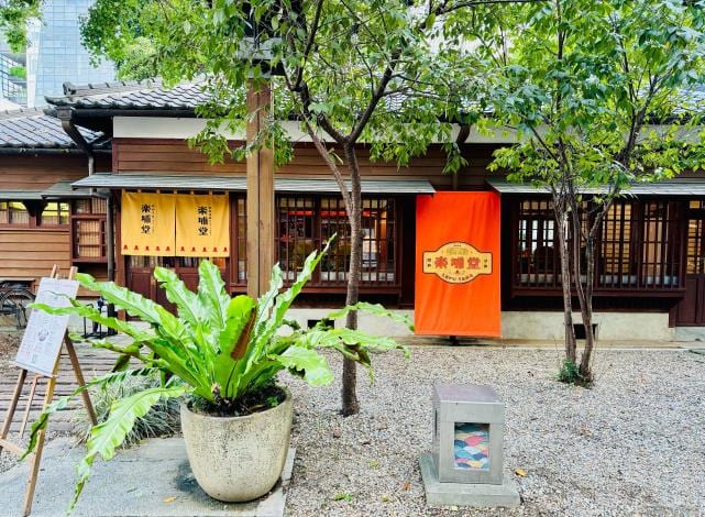A gravel courtyard before a Japanese-era timber house, with green foliage, a large bird’s-nest fern and an orange Lepu Tang banner.