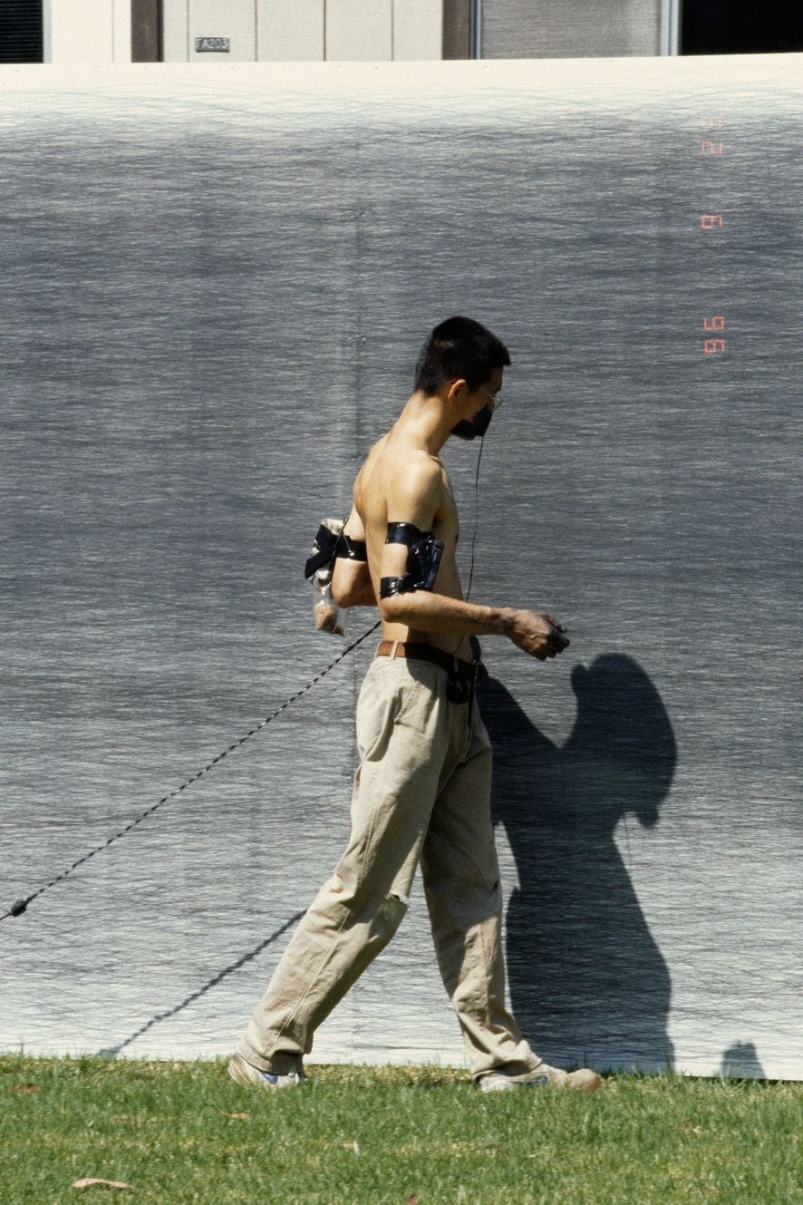 Shi Jin-Hua walks bare-chested beside a graphite-darkened wall with drawing equipment strapped to his body and a pencil line trailing behind him.