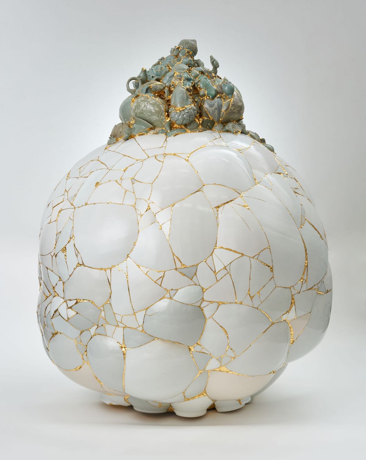 A broad rounded white ceramic body assembled from fragments, with celadon and decorated shards clustered densely at the top and every join drawn in gold.