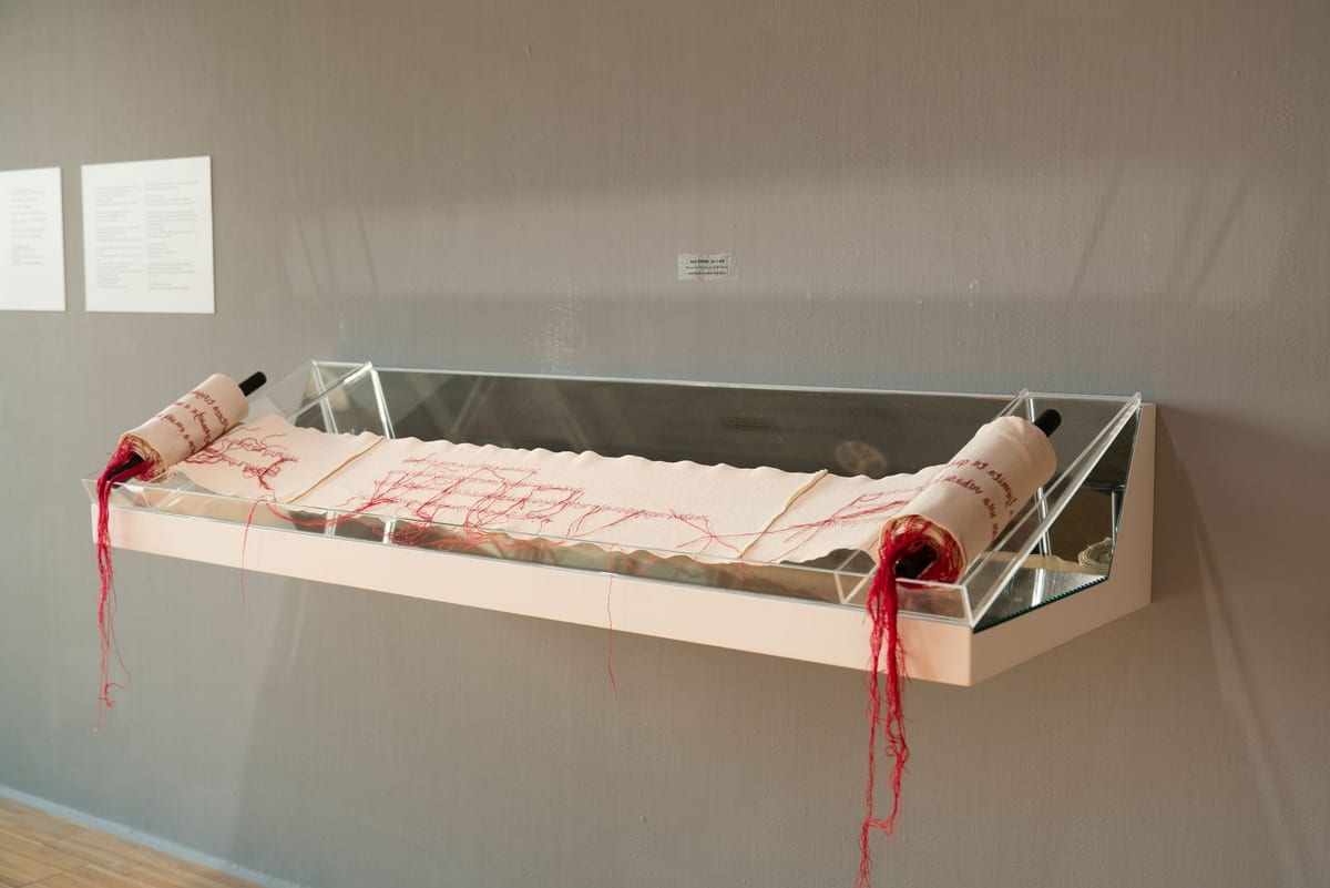A long pale cloth held half-unrolled on a clear wall support, red stitching running through Roman letters and other characters, with loose thread at both ends.