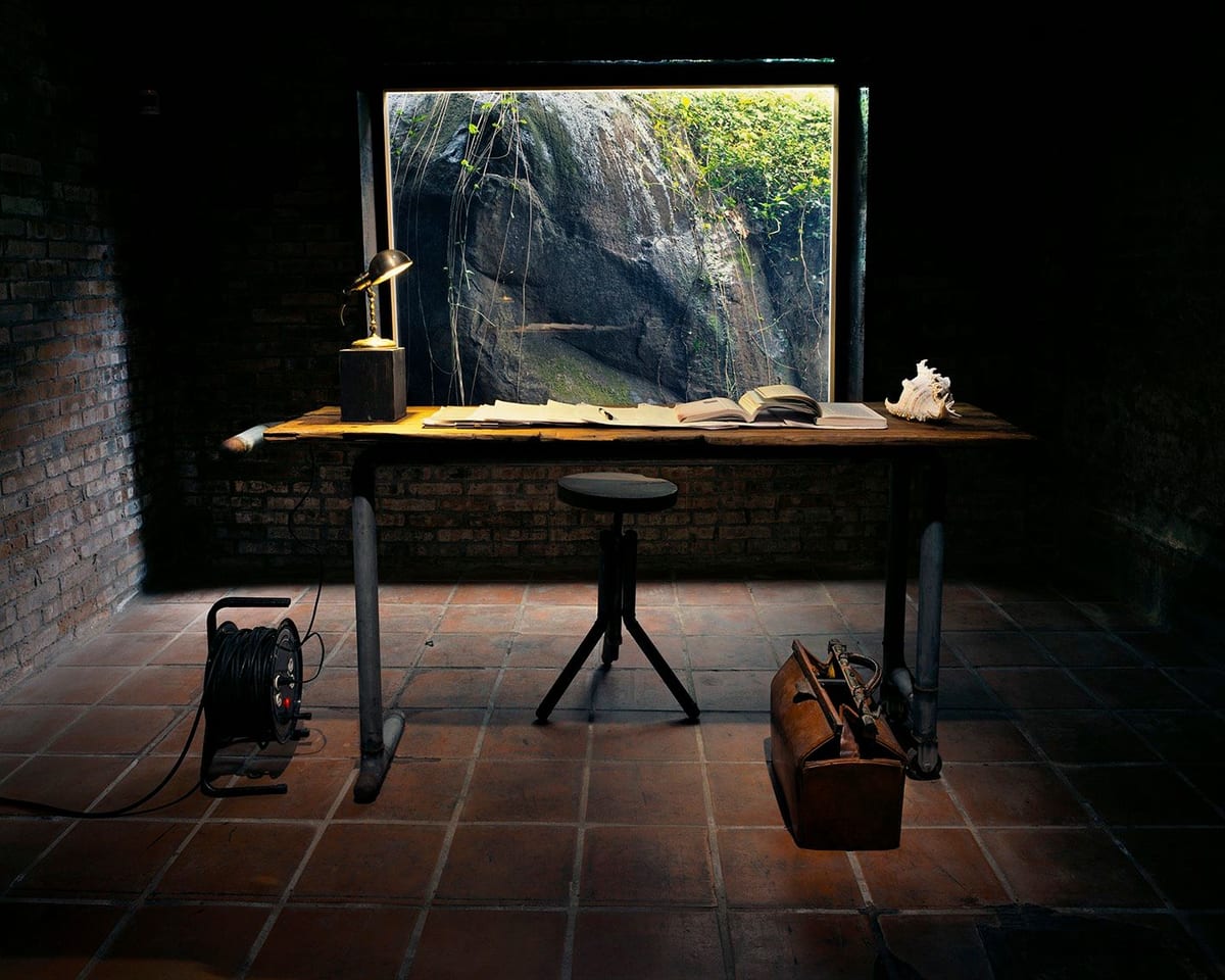 A dark brick room with a desk, stool, books, lamp, shell, cable reel and suitcase around a bright framed view of rock and vegetation.