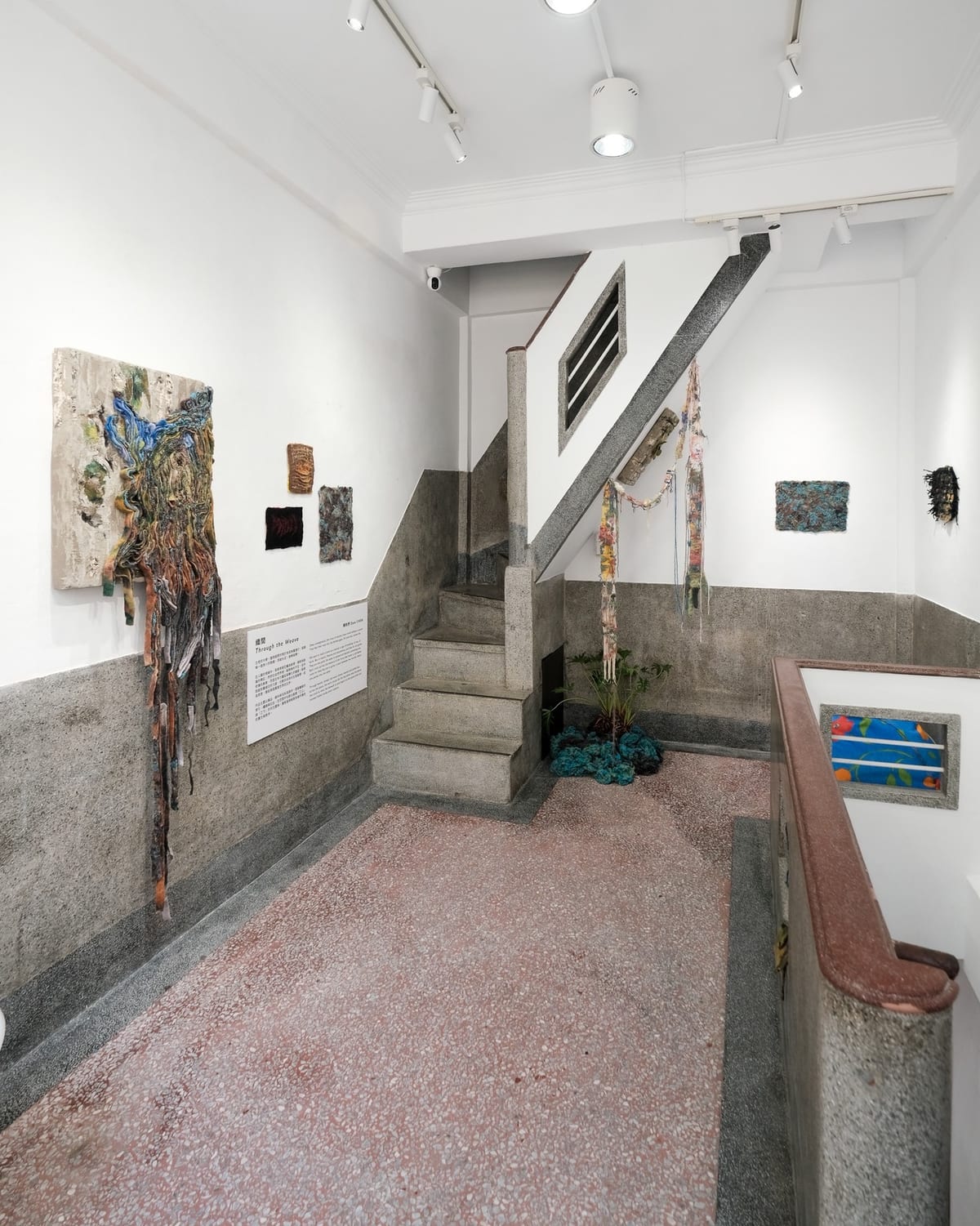 Coloured strands spill from a rough pale wall panel beside smaller textile pieces; a narrow terrazzo stair rises between the works, with long fibre pieces hanging above.