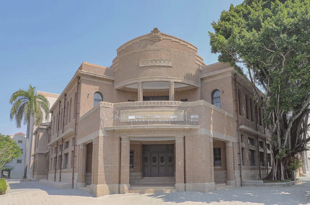 The warm-toned former police-station facade of Tainan Art Museum Building 1, with a stepped central gable and a large tree to the right.