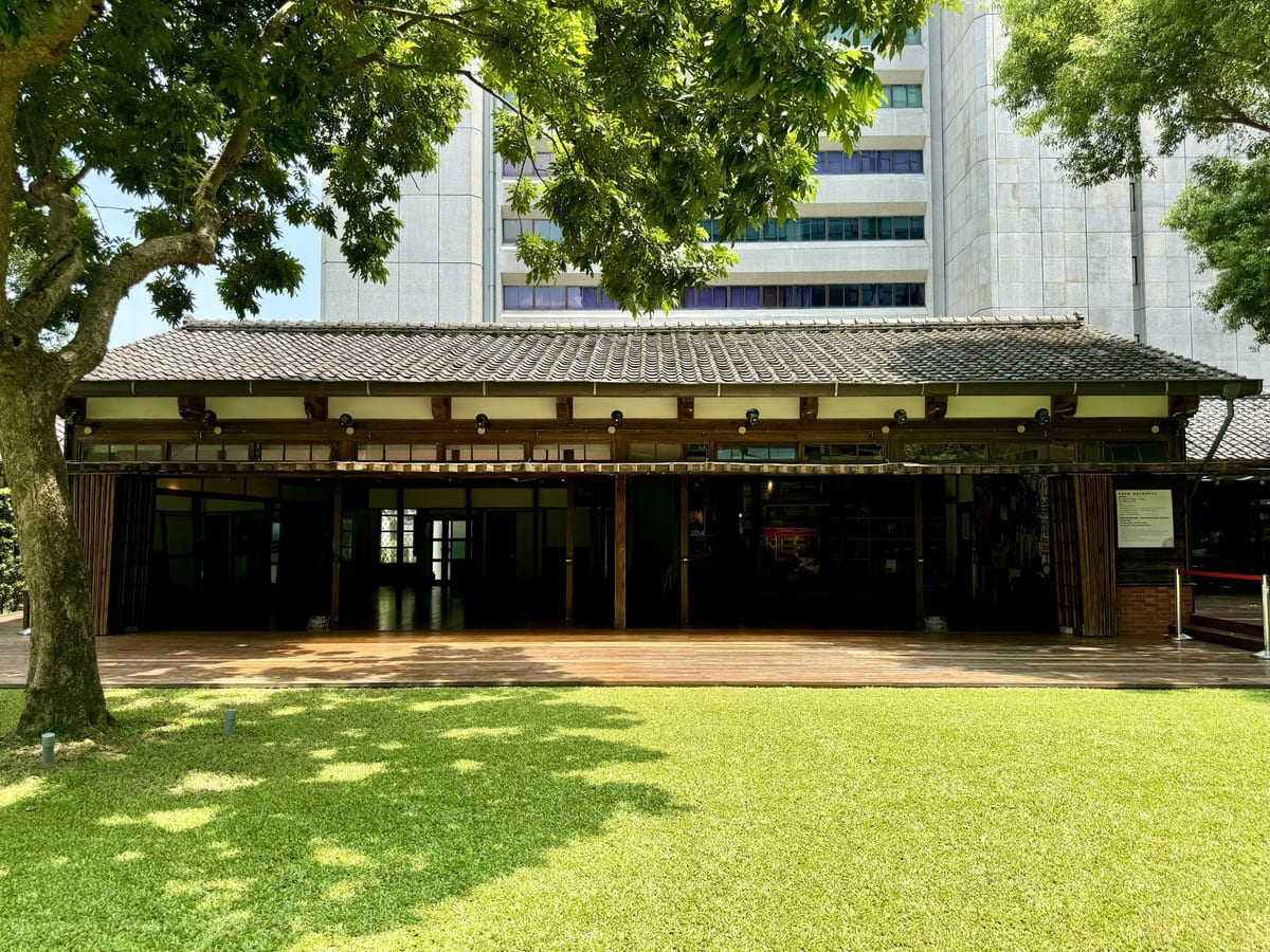 A single-storey Japanese-era wooden house behind trees and a lawn, with the high-rises of Zhongshan North Road behind it.