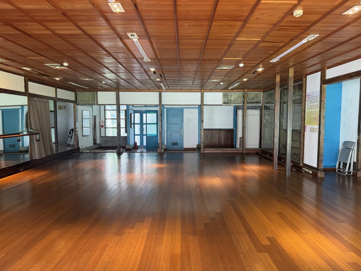 The interior of a wooden studio: a reflective sprung floor, windows running along three sides, dark timber beams overhead.