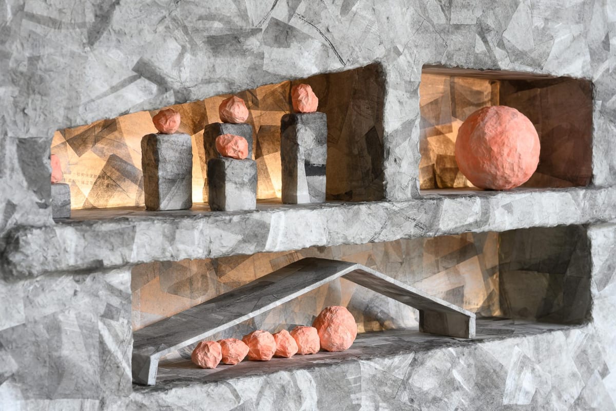 A grey paper structure has warmly lit recesses holding pink spheres, some on narrow blocks and others gathered behind a low paper rail.