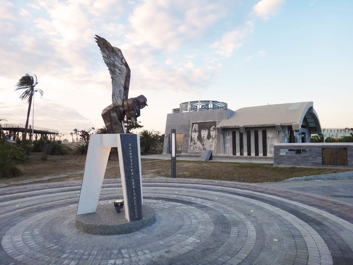In a seaside park, a winged dark figure stands on a plinth in front of the low theme museum under an evening sky.