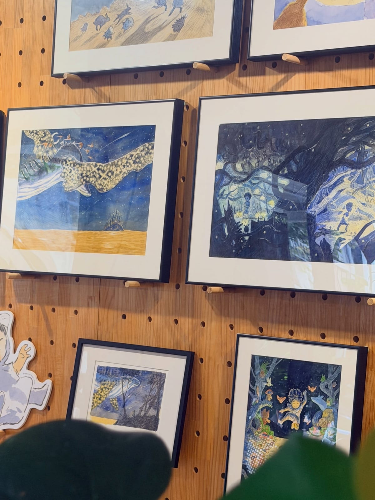 Framed blue-and-yellow picture-book originals hang on a wooden pegboard at cafe BOMBOM.