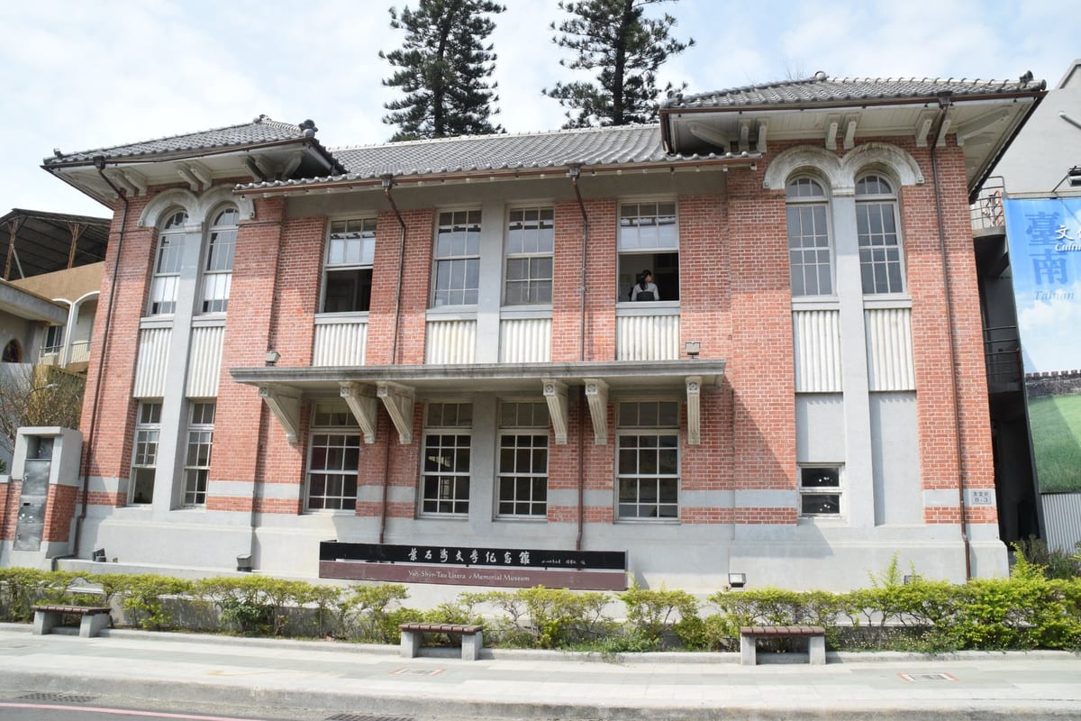 A two-storey brick and washed-stone building with steps and a signboard at the entrance, street trees in front.