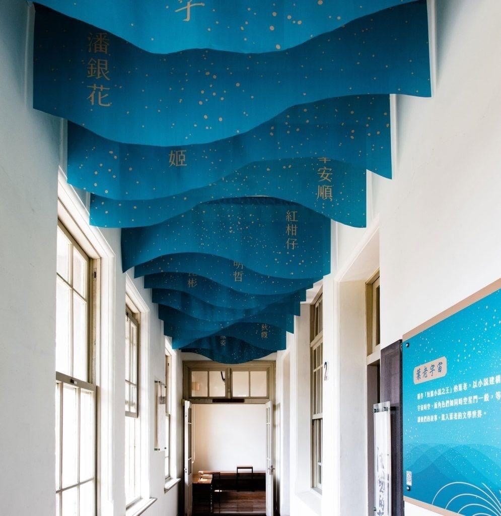 Blue wave-shaped panels printed with story titles hang from the ceiling above a wooden doorway into the next room.