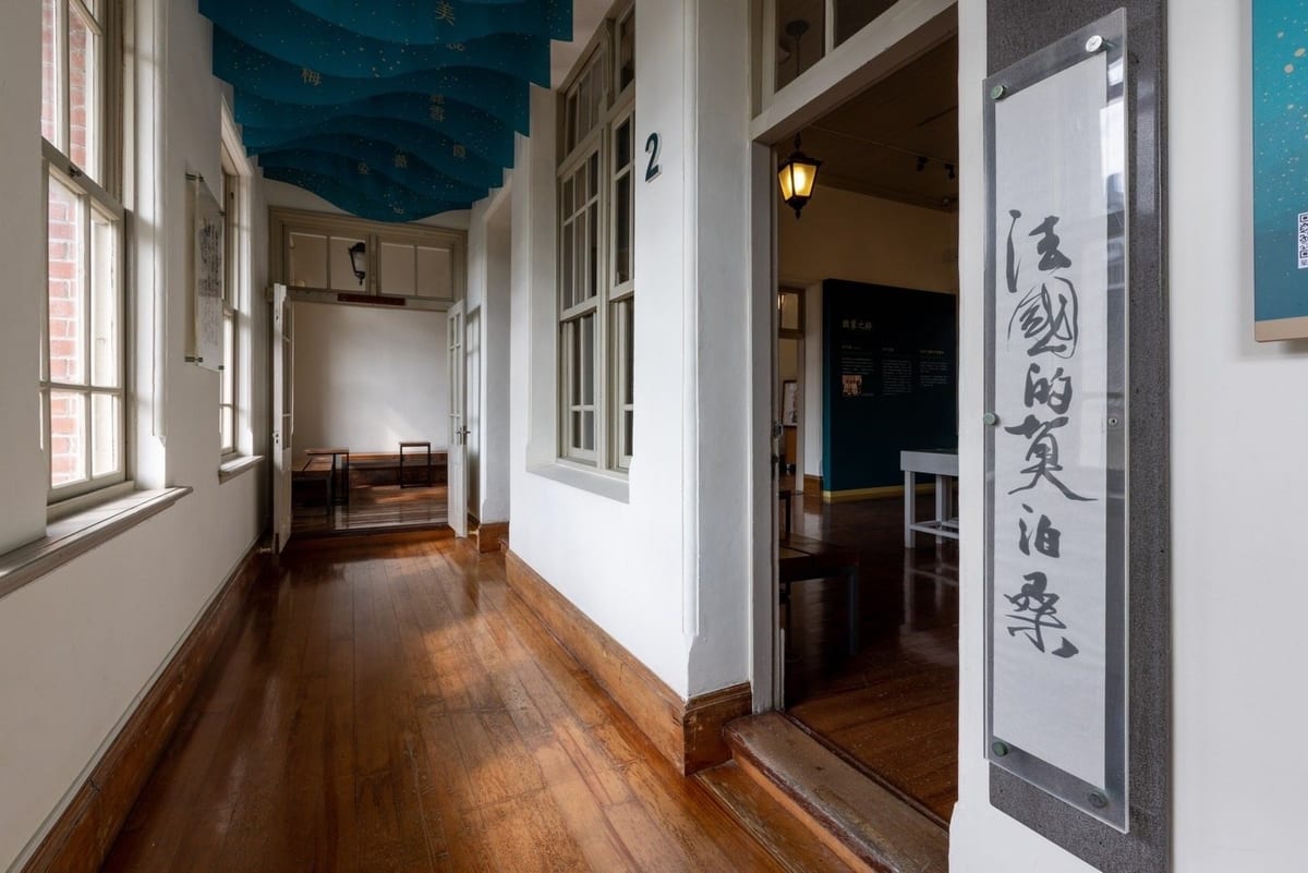 A wooden-floored corridor with white walls and old timber windows, calligraphy scrolls hung along it, opening into the next gallery.