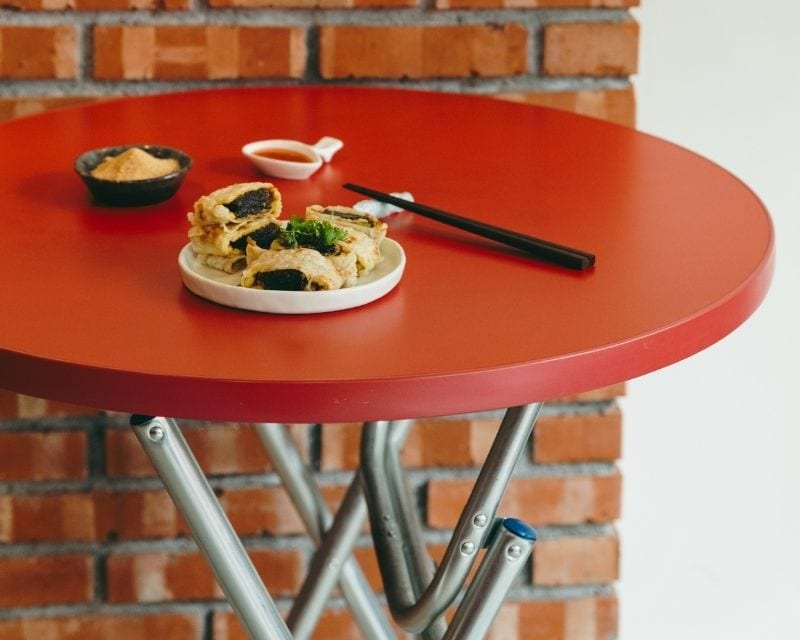A round red table on metal legs in front of a brick wall. A white plate holds cut fried rolls with a dark filling and a sprig of herbs, beside a small bowl of brown powder, a dish of sauce and a pair of chopsticks.