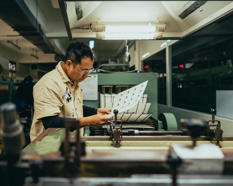 A man in glasses and a khaki work shirt stands at a green printing machine, holding up a sheet printed with rows of small blue-and-white motifs; metal machine parts fill the foreground.