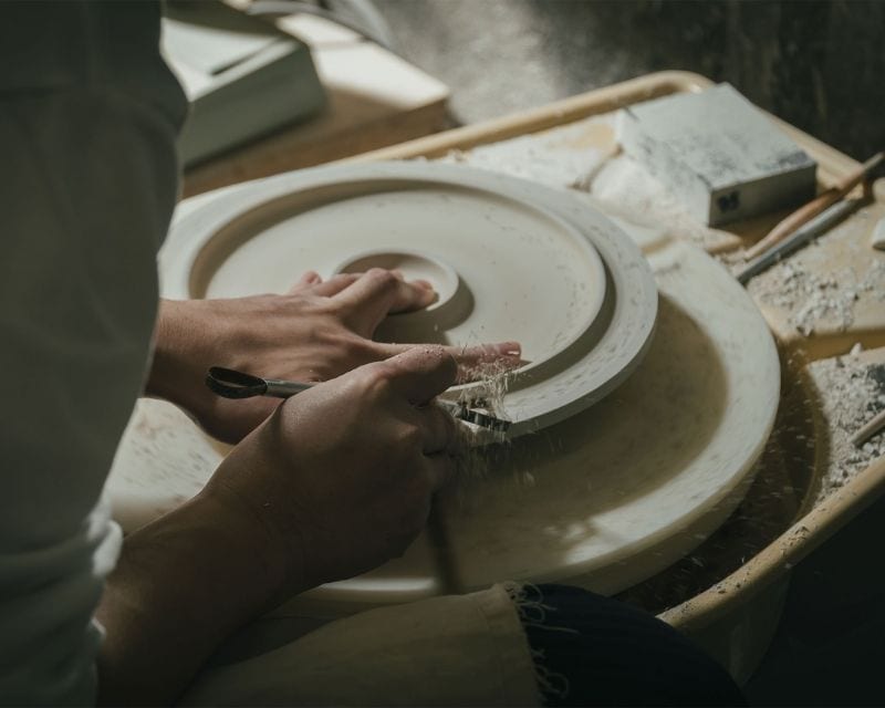 Close-up: one hand holds a pale, upturned unfired bowl on a turning wheel while the other trims its foot with a metal tool, sending curls of clay flying; scraps and tools lie on the tray beside it.
