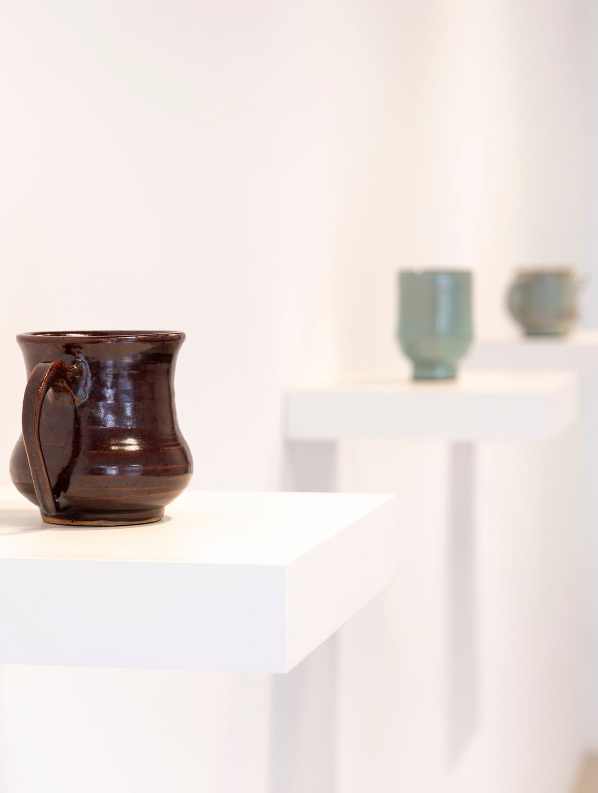 A brown glazed vessel stands on a white plinth, with two pale green vessels blurred behind it.