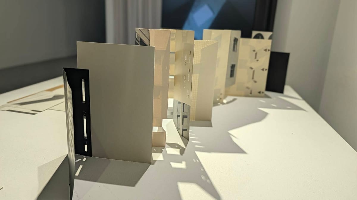 Pale folded pages stand in small architectural compartments on a white table; dark panels and cut-outs cast long sunlit shadows.