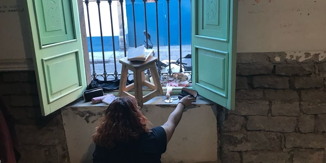 Green shutters open around a window. Papers rest on a wooden stool while a person reaches towards scattered objects; a blue door stands beyond the iron bars.
