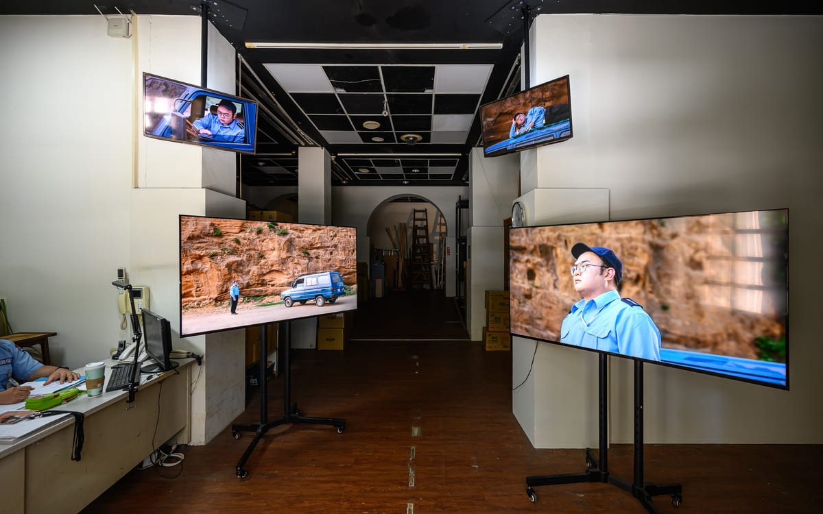 Four monitors around a corridor show a uniformed man and a blue van beside rock. A staffed desk is visible at the left edge.