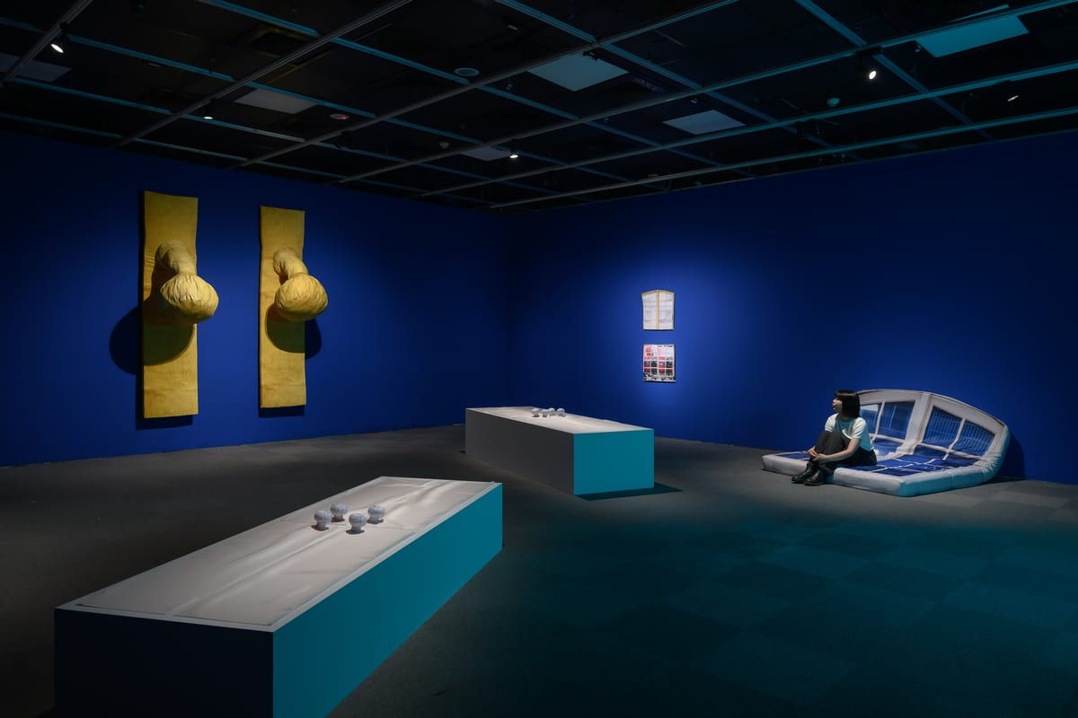 A cobalt-blue room contains two padded yellow door-handle shapes, long plinths with small rounded objects, and a white upholstered window-seat form occupied by a visitor.