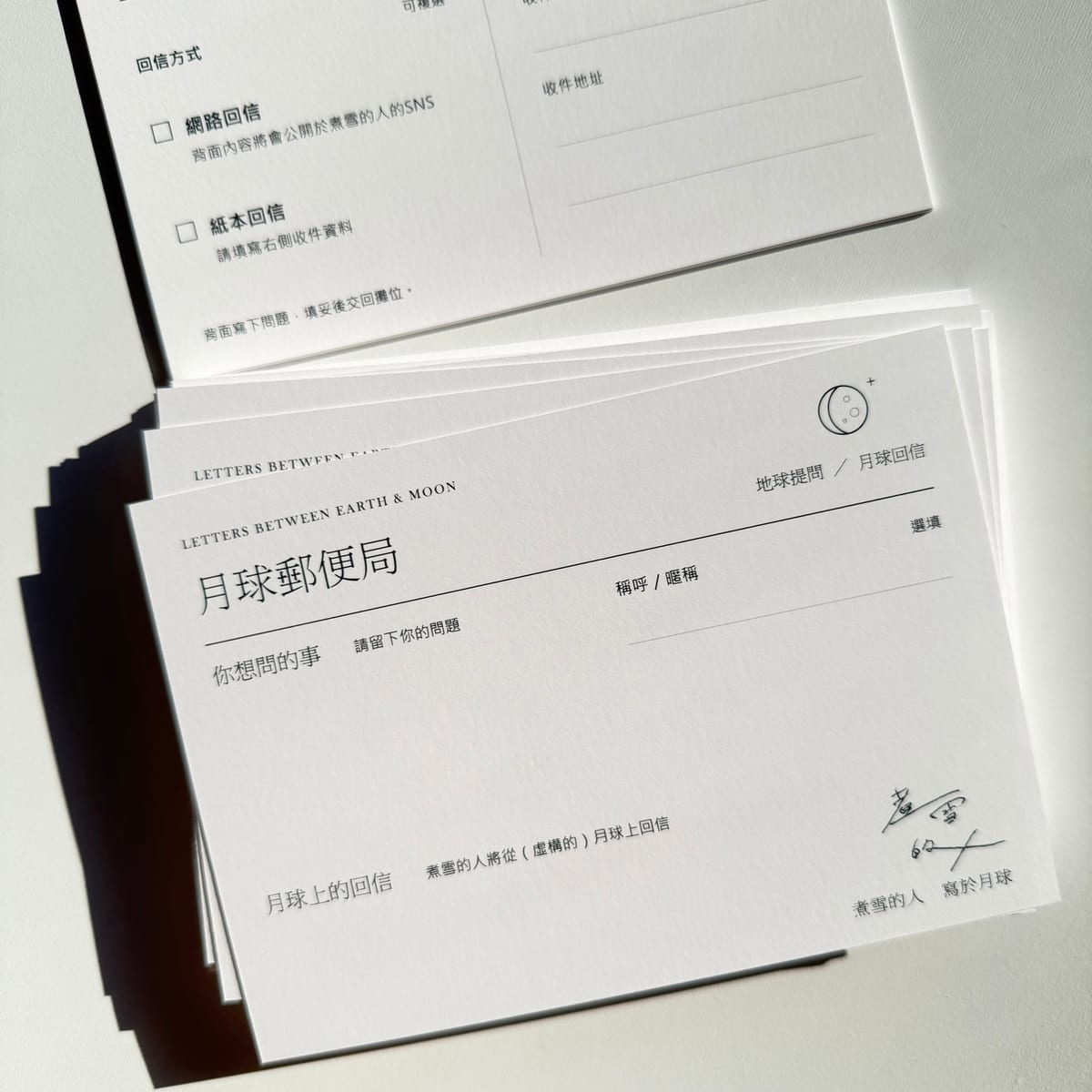 Moon Post Office postcards lie stacked on a pale surface, with spaces for a question from Earth and a reply from the moon; the reverse offers online or postal replies.