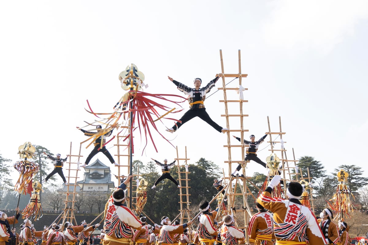Performers in black-and-white coats extend their limbs from tall ladders while ground teams hold them steady; red and gold streamers rise in front of a castle.