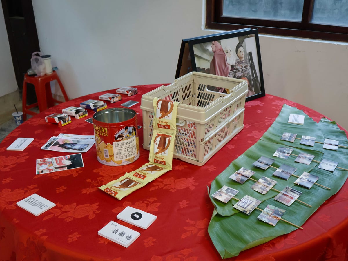 Small photographs on sticks cross a broad green leaf on a red tablecloth, beside a biscuit tin, hanging coffee packets, a pale basket and a framed photograph.