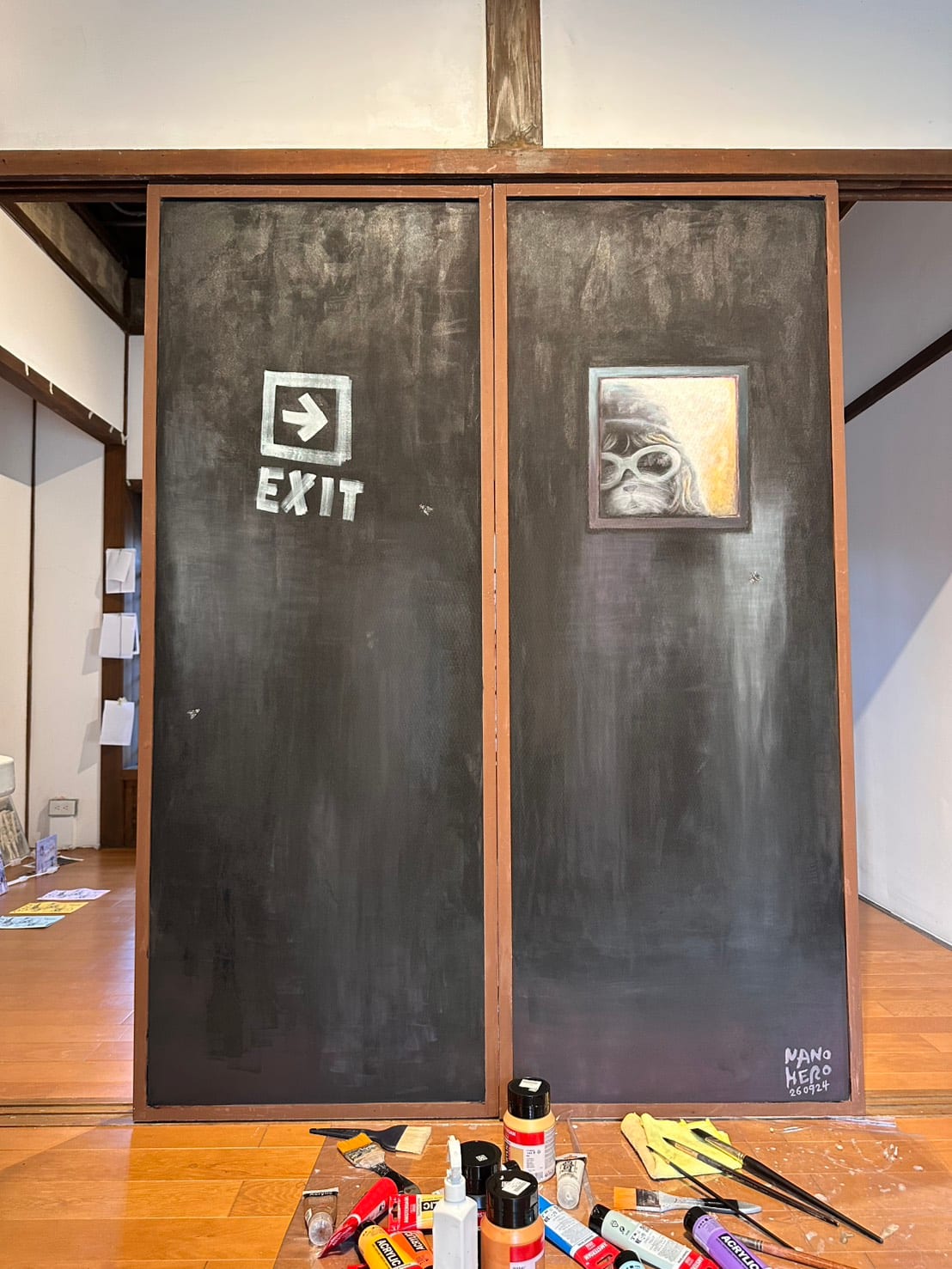 The reverse of the two panels is painted dark grey, with a white EXIT arrow on the left and a small painted portrait of Toby on the right. Brushes and paint bottles sit below.