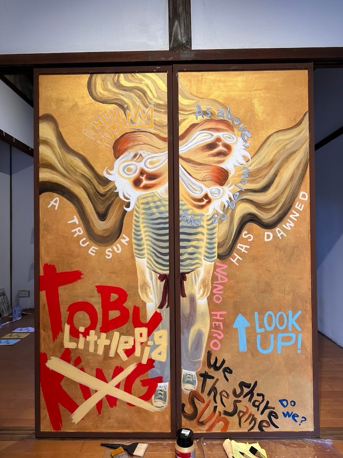 Two wooden door panels painted gold carry several distorted, goggle-wearing Toby figures and hand-painted English text. Paint bottles and brushes sit on the floor in front.