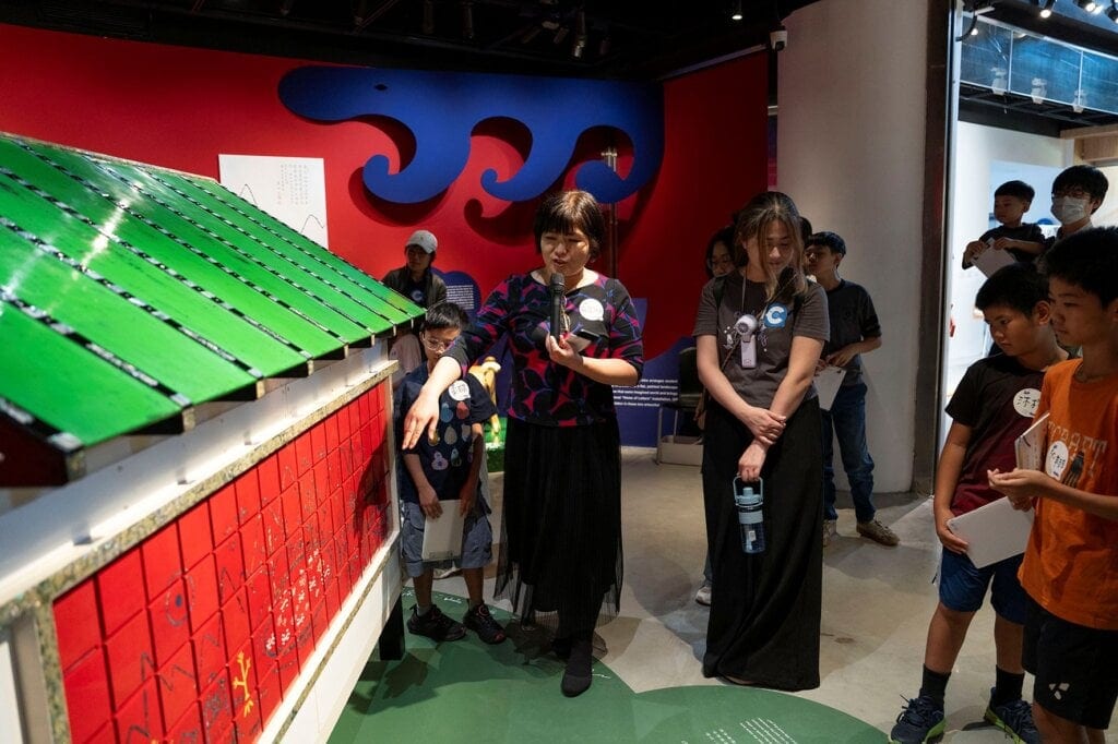 Song Joo Yean introduces a green-roofed house with red puzzle panels to families; a blue wave hangs on the red wall behind her.
