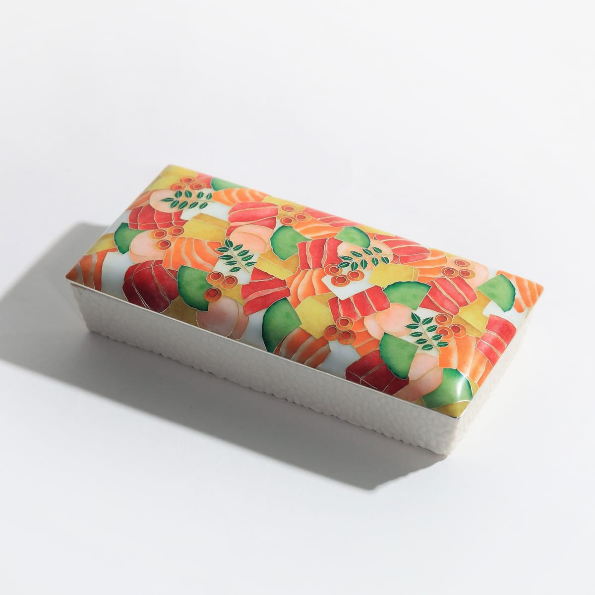 Fish, prawns, roe and green leaves decorate a dark rectangular box with a glossy surface, photographed against grey.