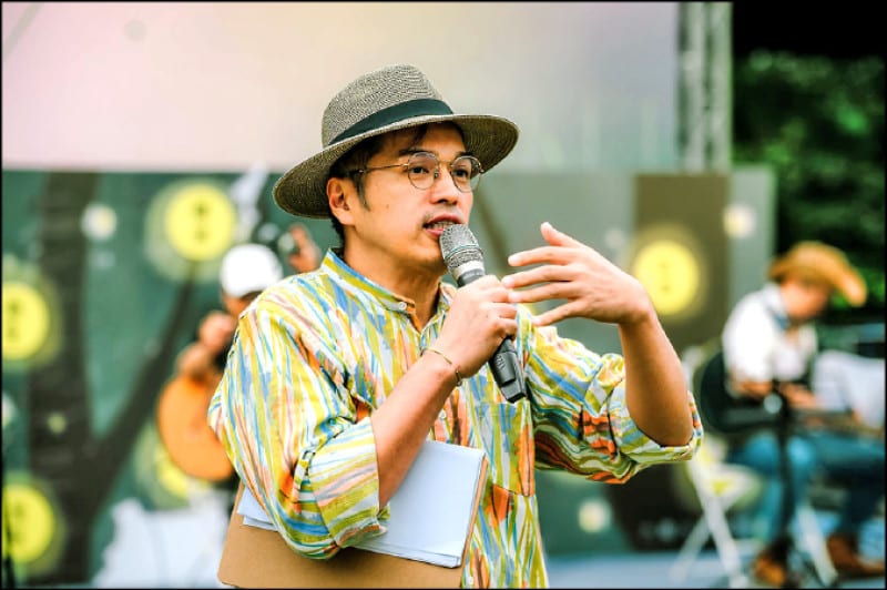 In a bright striped shirt and hat, Lin Qing-sheng speaks into a handheld microphone inside the bookstore, with shelves and the long table behind him.