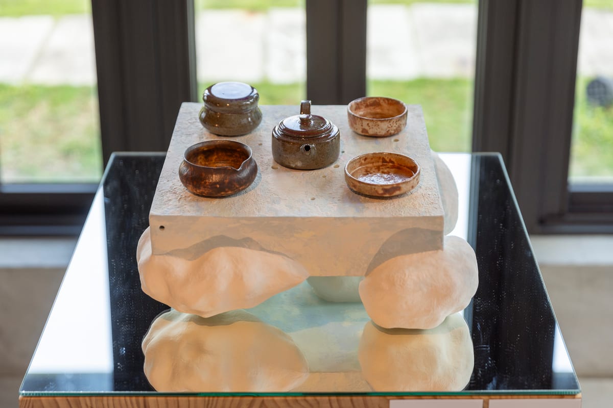 Five brown and amber tea vessels on a pale cloudlike ceramic support, reflected in glass below.