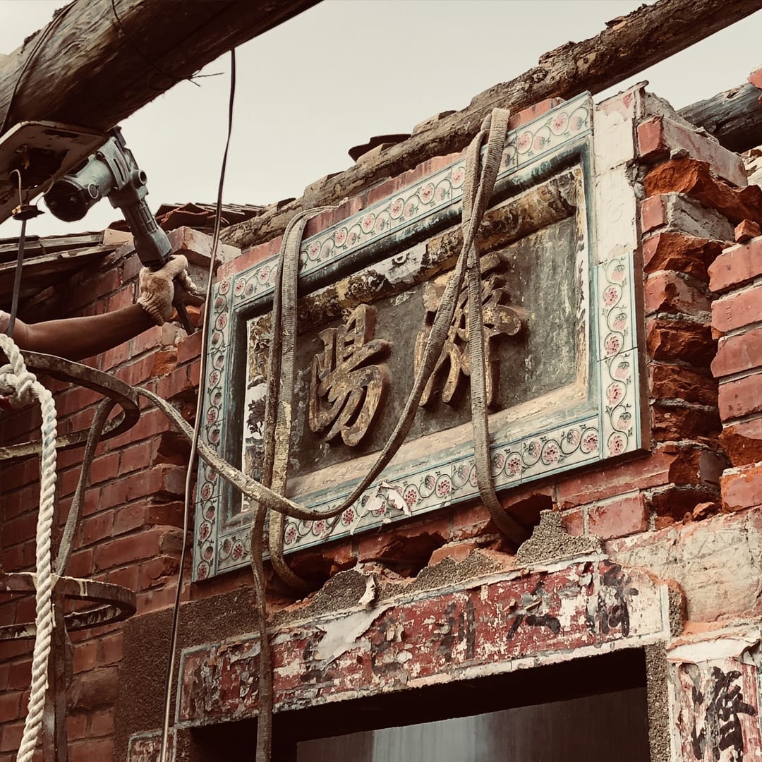 Two thick ropes hang in front of a plaque bearing the characters 濟陽, framed by pink flower tiles, while a worker tackles the surrounding brickwork.