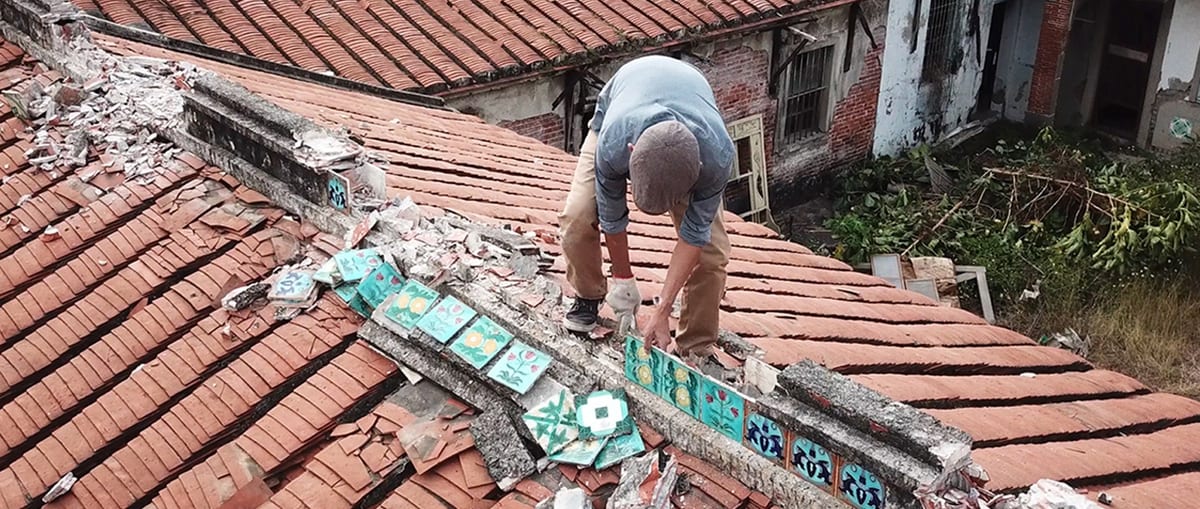 A worker removes turquoise flower tiles from broken masonry above a red-tiled roof; loose tiles lie beside his feet.