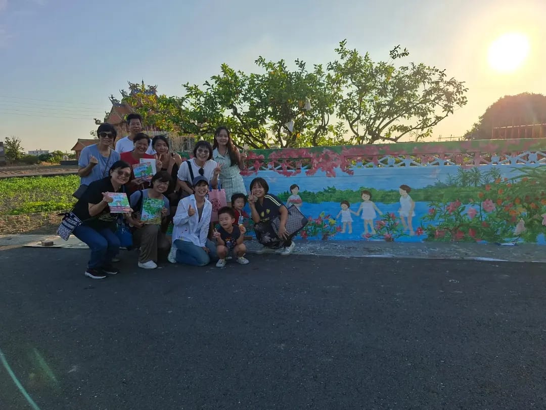 Visitors and children pose beside a low painted wall at sunset in Xuejia. Blue water, people, flowers and birds run along the wall, with trees and a temple behind.