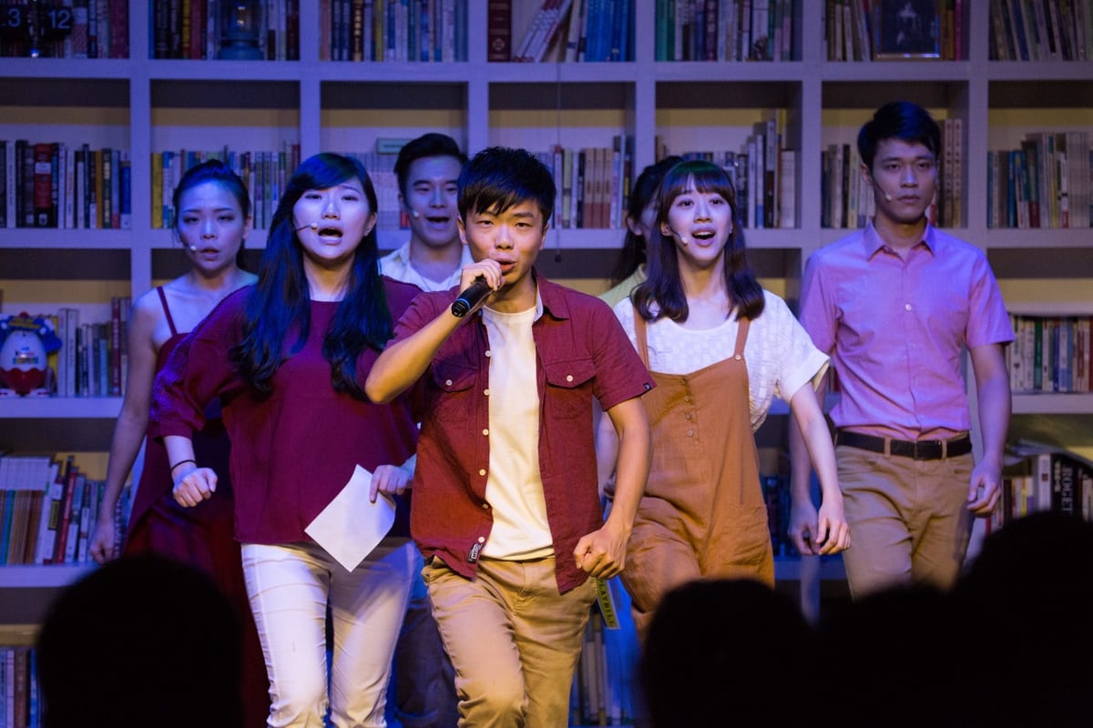 Six A劇團 performers sing in front of bookshelves under blue and purple stage lighting.