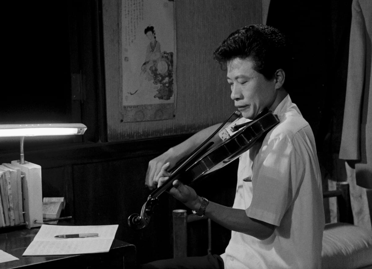 A black-and-white film still shows a man playing a violin beside a desk, papers and a lamp.