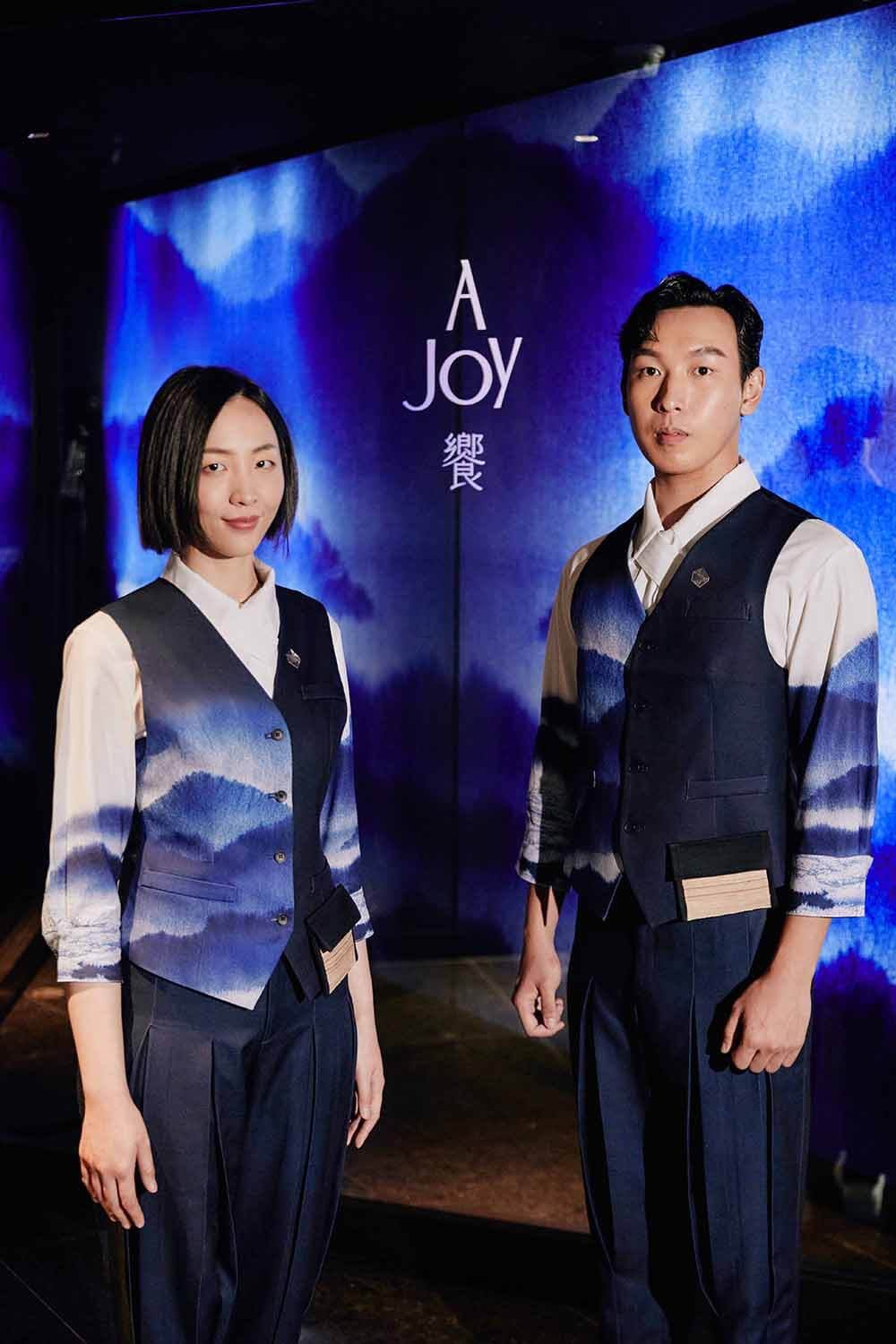 Two people wear white shirts and navy waistcoats against a blue backdrop. Blue-and-white mountain landscapes cross the sleeves and torsos, with small badges on the waistcoats.