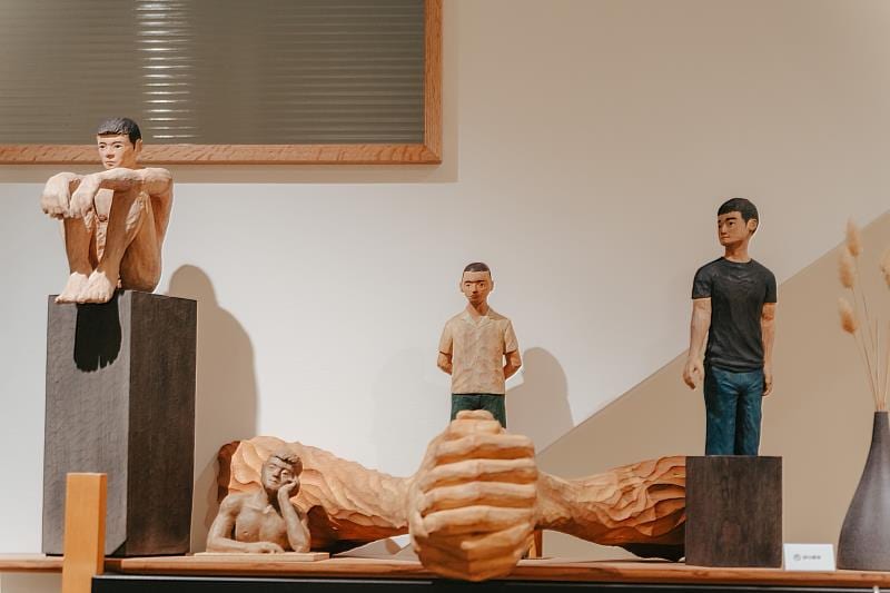 Four carved wooden people around an enormous wooden hand: one hugs his knees on a tall dark plinth, another rests his head beside the hand, and two stand behind in pale and black shirts.