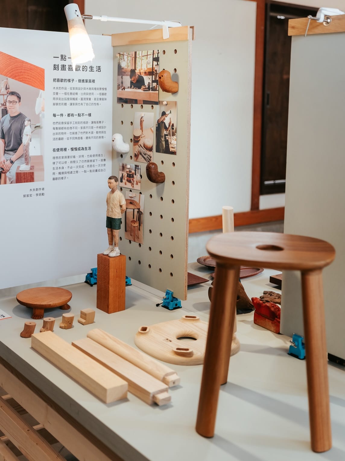 A three-legged wooden stool with a central hole, separate stool parts, a carved figure and bird hooks stand before white pegboard; the panel at left introduces the two makers.