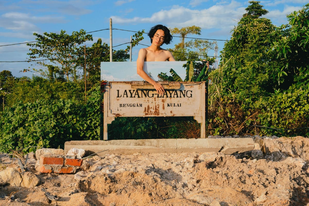 Xhin Toh stands bare-chested in a green landscape with broken ground, holding a photograph of a weathered LAYANG-LAYANG station sign across his body.