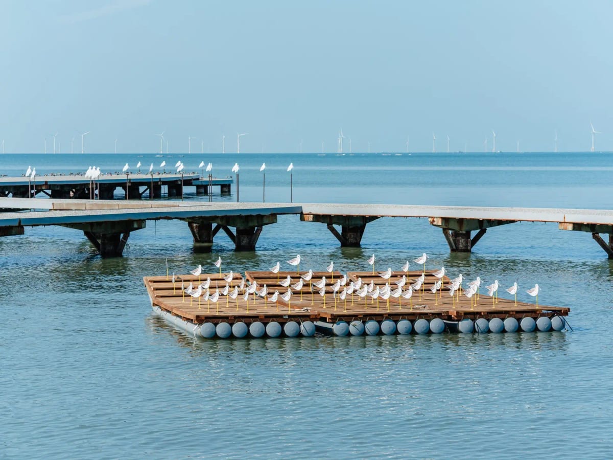 White bird-shaped wind vanes stand on a raft, with the crab boardwalk and distant wind turbines behind them.