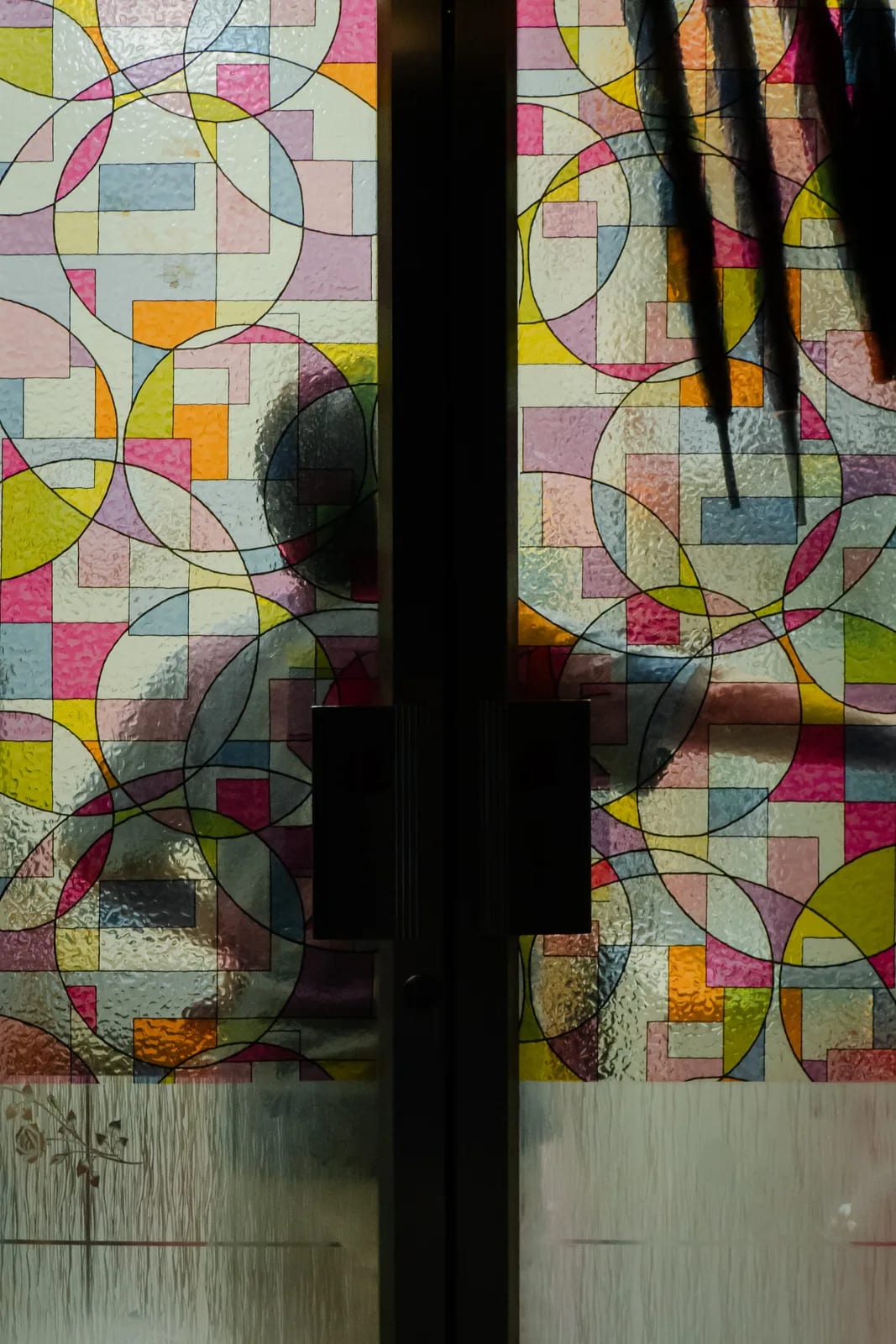 A partly shadowed doorway has textured glass covered with interlocking pink, yellow, green and pale-blue shapes.