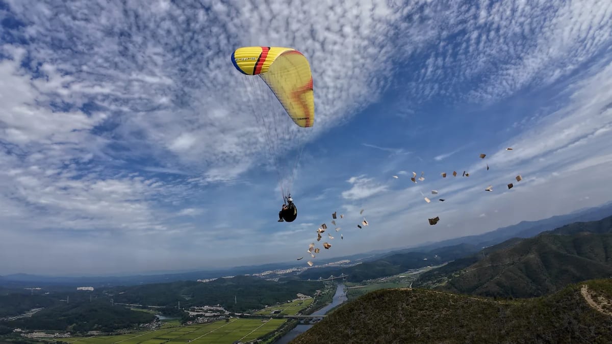A paraglider floats over a river valley while small pieces of paper scatter through the air behind the performer.