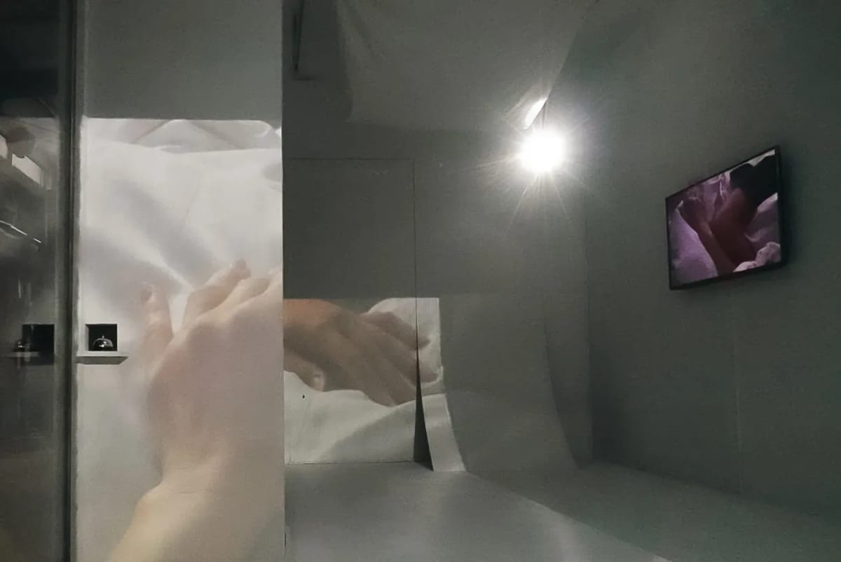 Moving images of hands are projected onto freestanding white surfaces in a dim room, with another image playing on a wall-mounted screen.
