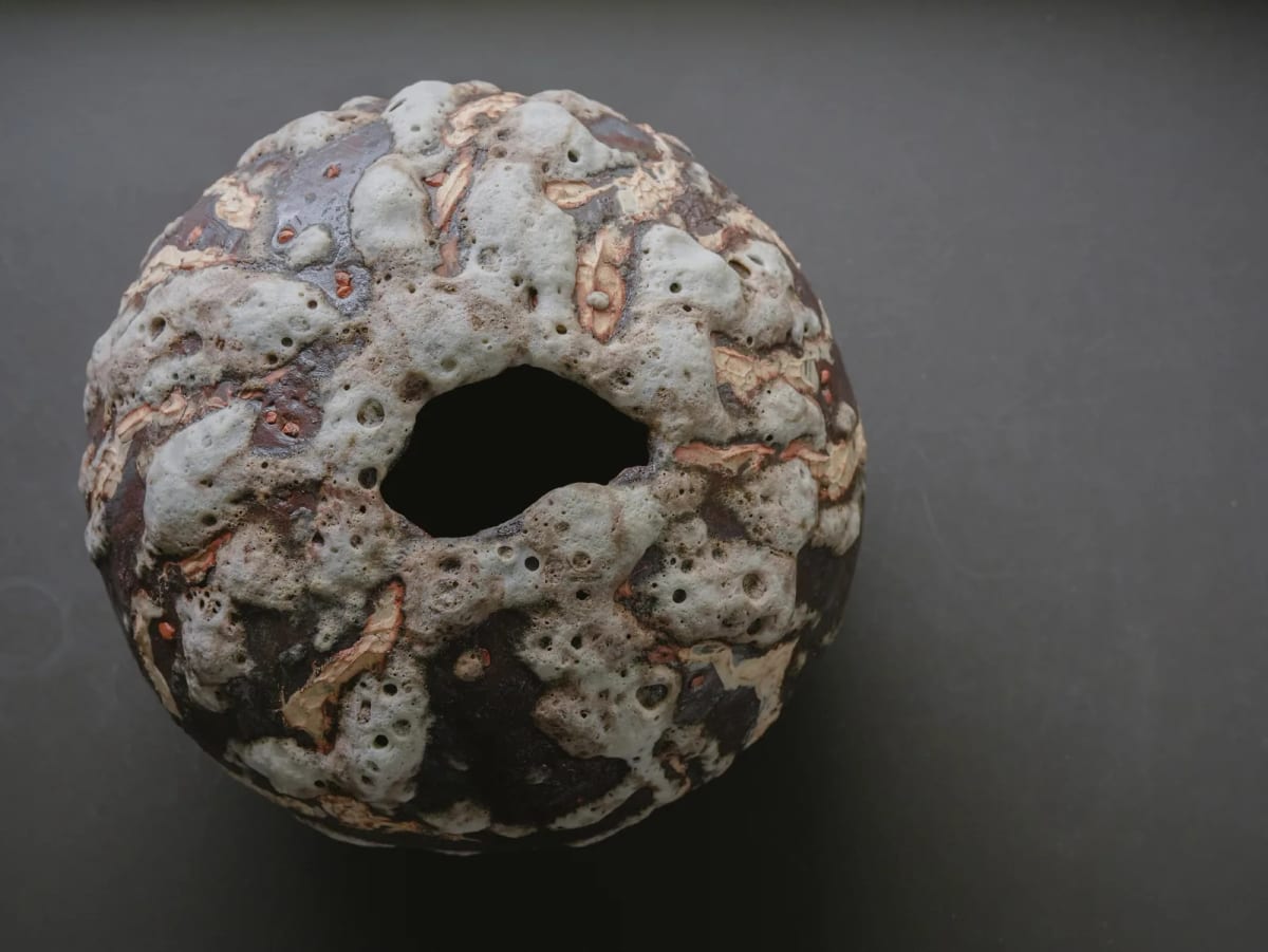 A rounded ceramic form has a dark opening, bands of clay and a rough, pale, pitted surface.